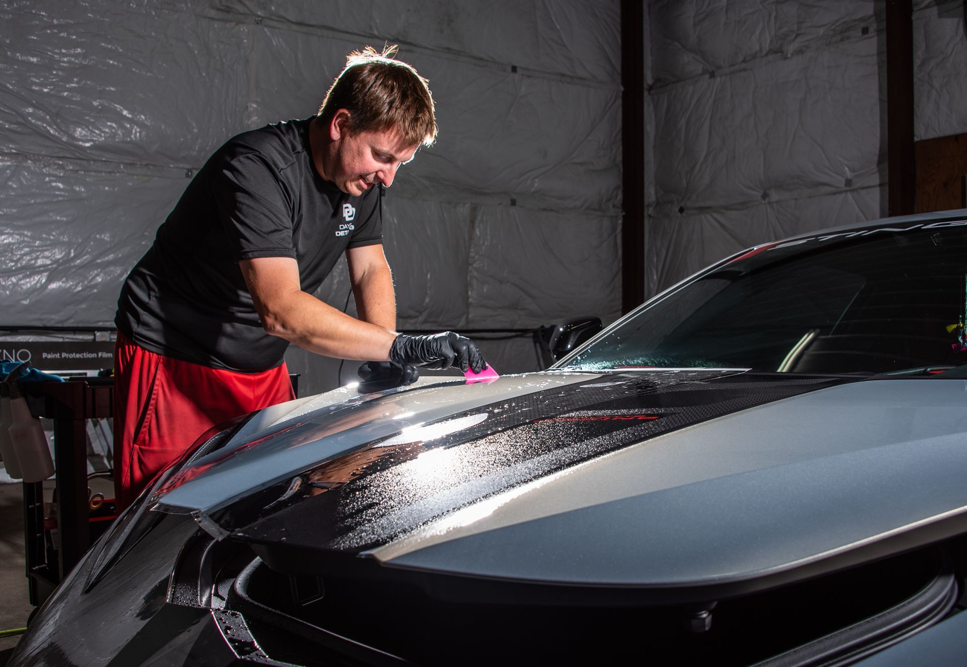 Man in black shirt washing a two-tone car hood inside a garage, using a pink pad.