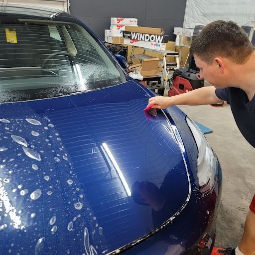 Person applying protective film to a blue car's hood in a workshop.