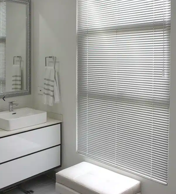 Baño moderno con tocador blanco, espejo de pared, persianas y un banco blanco junto a la ventana.