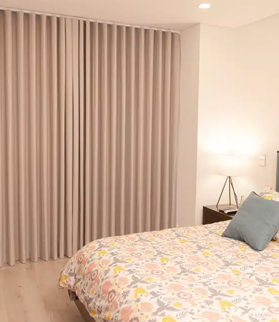 Habitación luminosa con cortinas beige, una cama con ropa de cama estampada con flores y una lámpara de noche encendida.