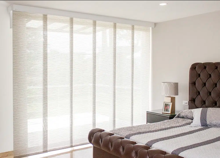 Habitación luminosa con cama capitoné, lámpara de noche y persianas altas que dejan entrar una luz natural suave.