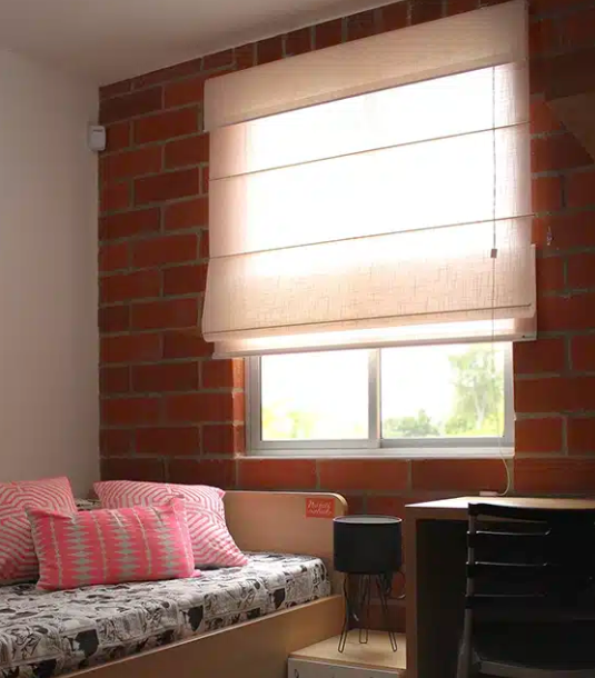 Habitación pequeña con una cama junto a una pared de ladrillos, almohadas rosas y una ventana con una persiana romana beige.