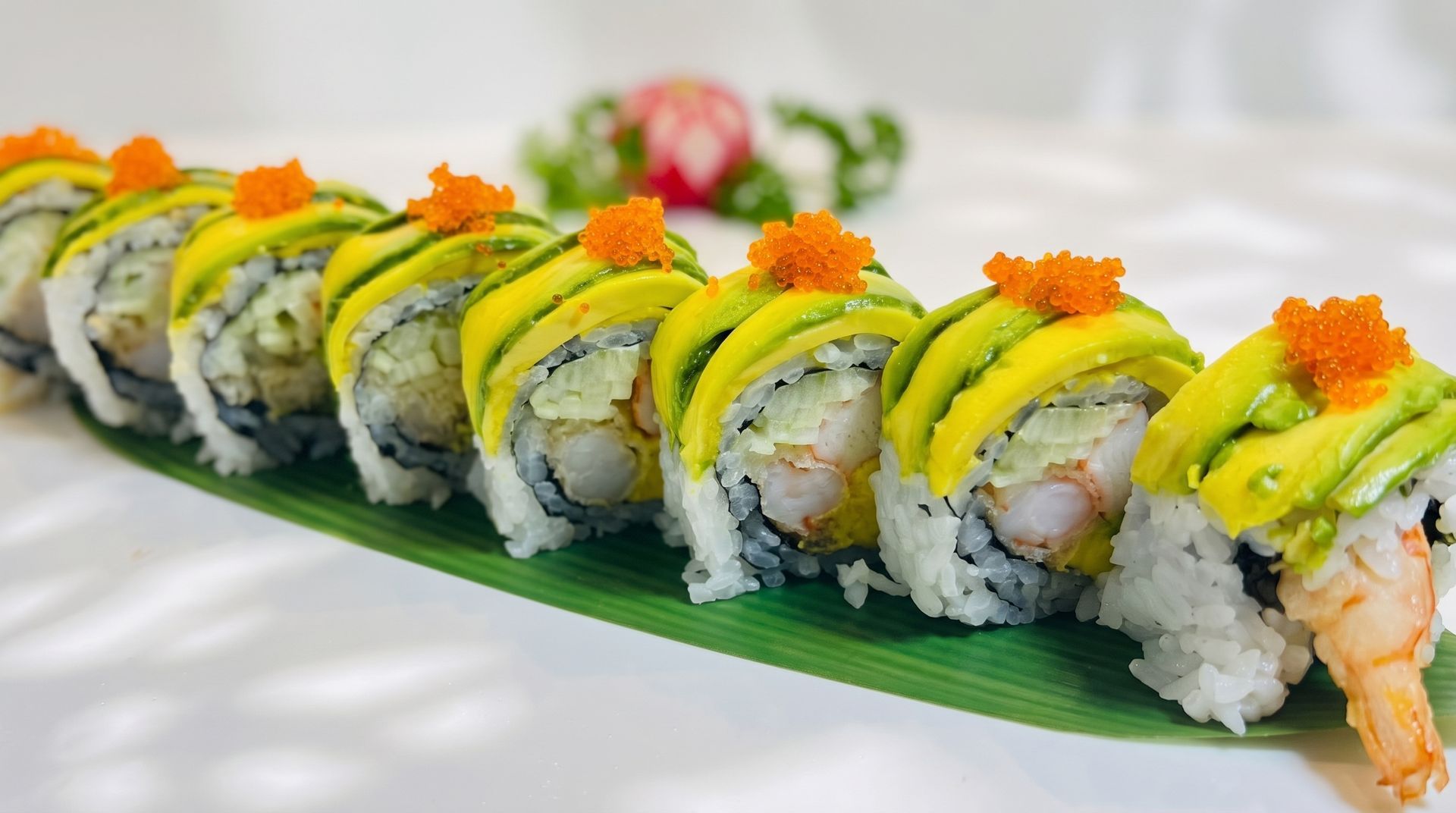 Sushi rolls topped with avocado and orange roe on a green serving leaf, with wasabi and pickled ginger nearby