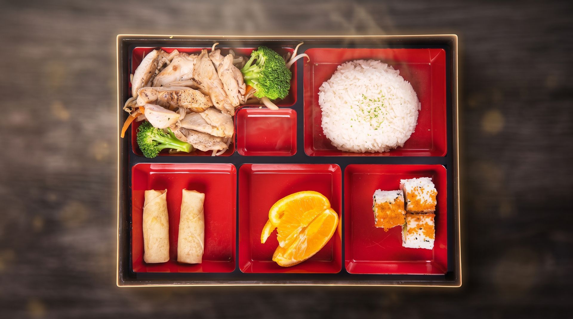 Bento box with rice, sliced meat, spring rolls, pickled vegetables, and orange slices in red compartments