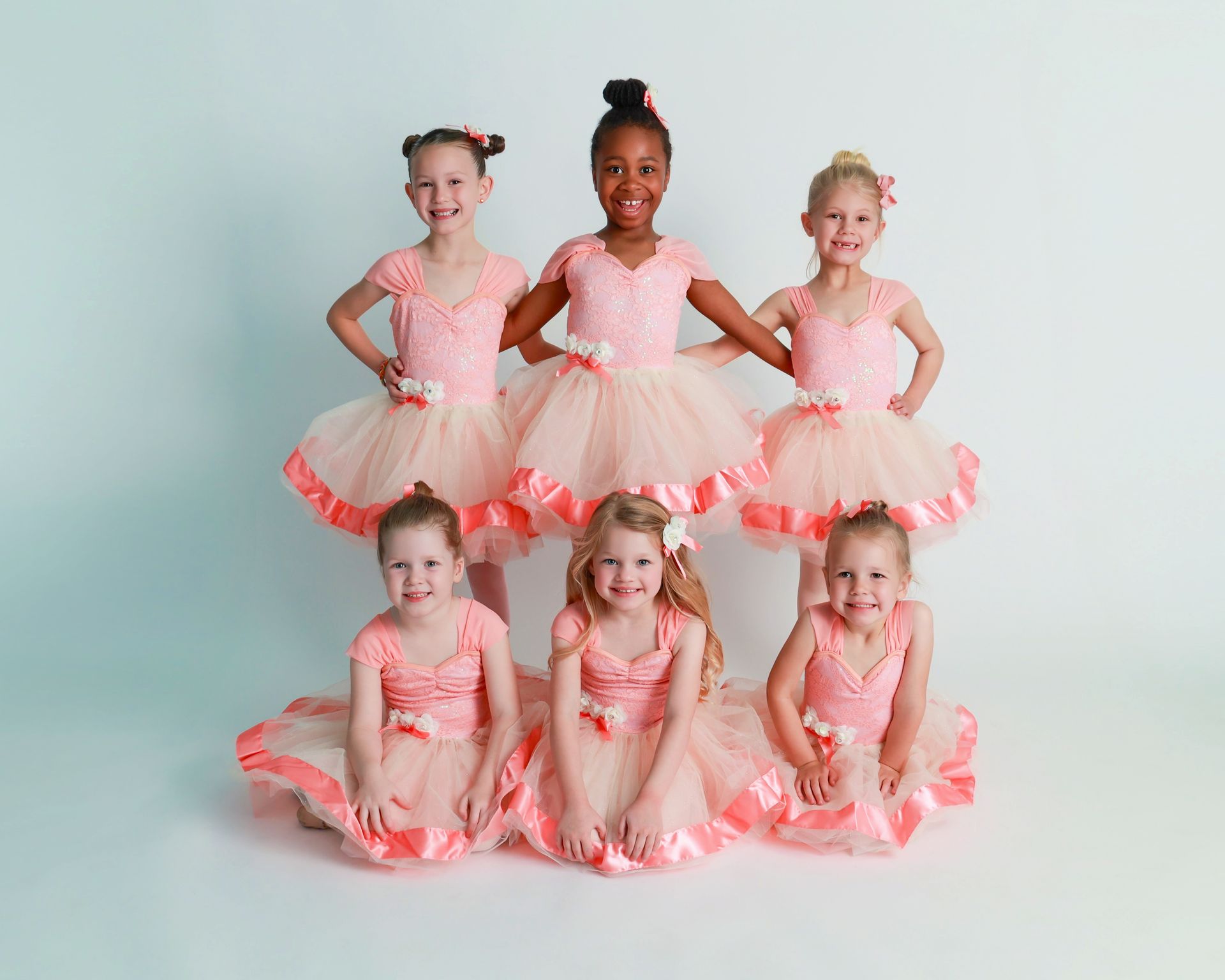 Six children in pink ballet tutus posing in two rows against a plain light background