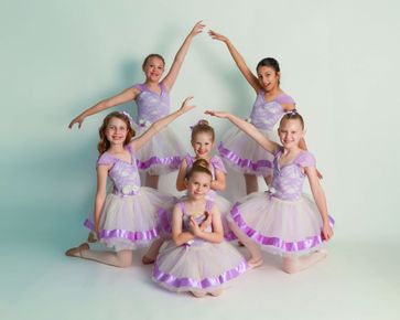 Six young ballet dancers in lavender tutus pose with raised arms in a studio.