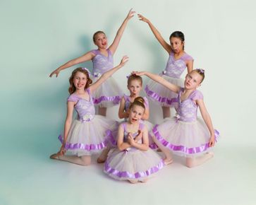 Five dancers in purple tutus pose in a studio, arms raised in a synchronized ballet formation.