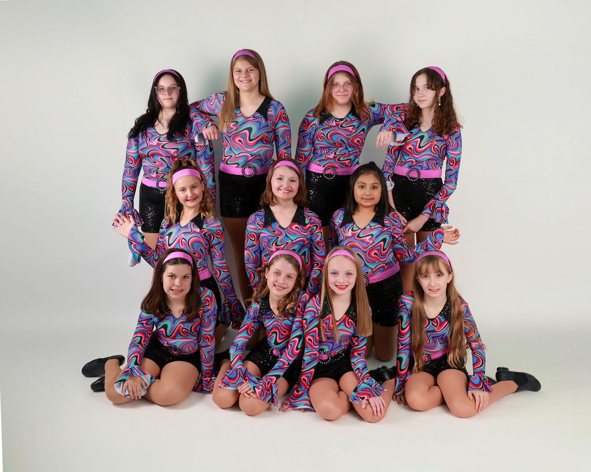 Group of ten dancers in colorful pink, blue, and black costumes posing against a white backdrop