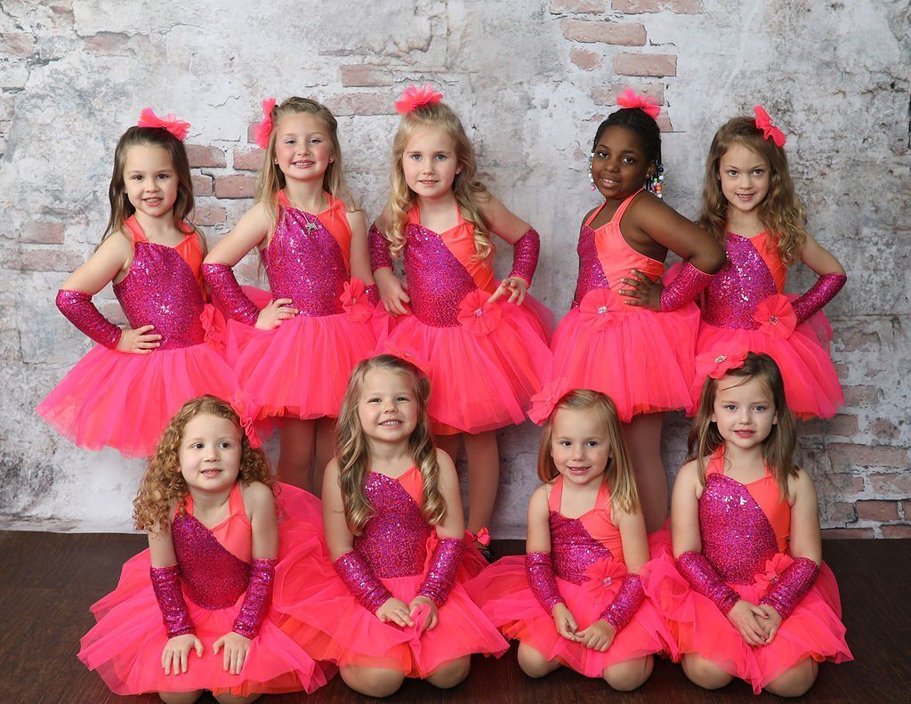 Nine young girls in pink dance costumes pose in front of a brick wall.