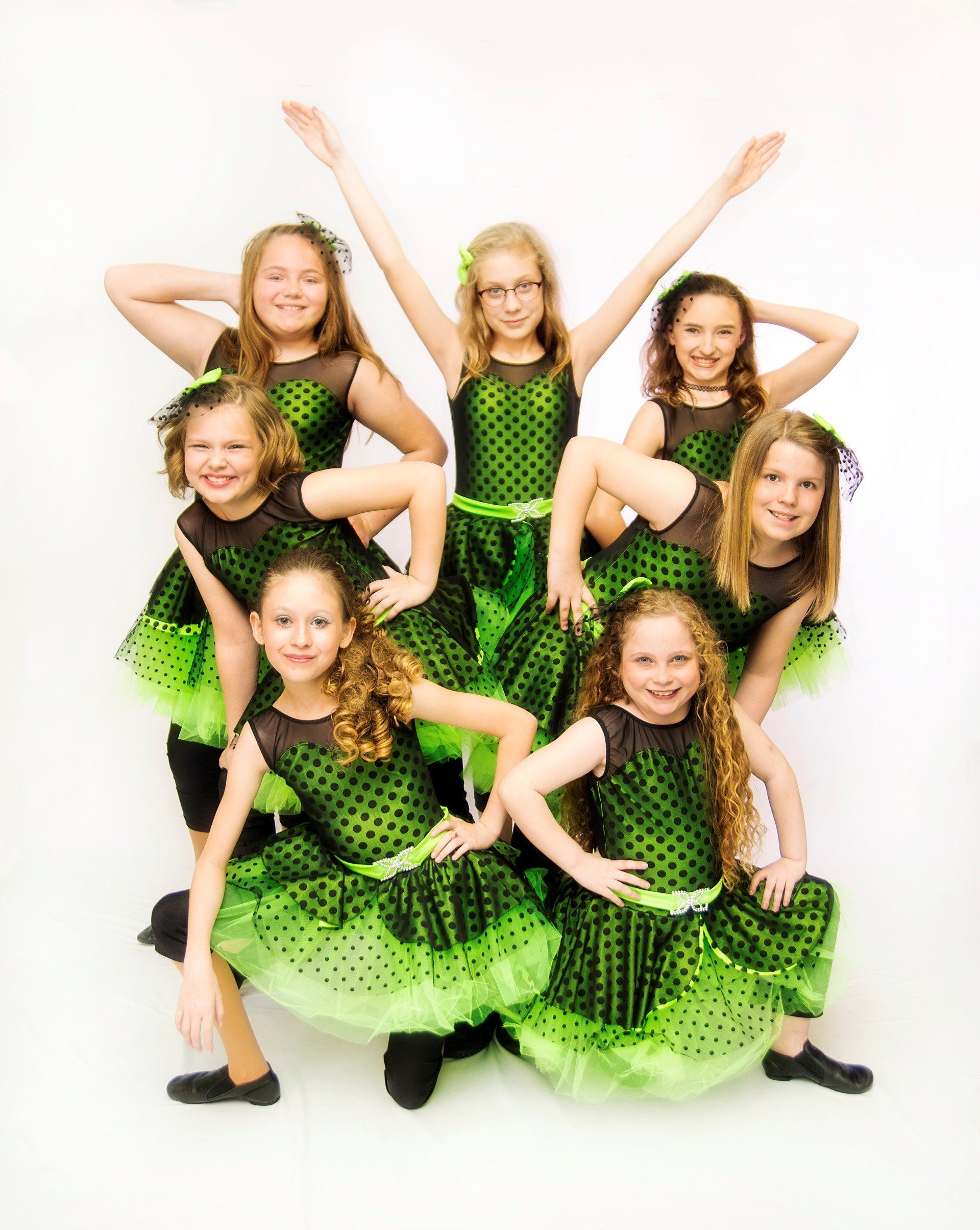 Seven young girls in green and black polka dot dance costumes pose in front of a white background.
