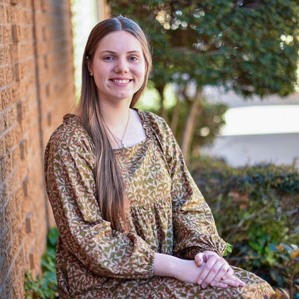 Woman sitting outdoors beside a brick wall, smiling in a patterned dress.