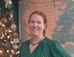 Smiling person in a green patterned top posing indoors beside a decorated Christmas tree