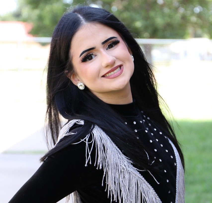 Smiling woman with long dark hair in a black outfit with silver fringe, standing outdoors in a park