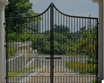 A wrought iron gate is open to a driveway with trees in the background.