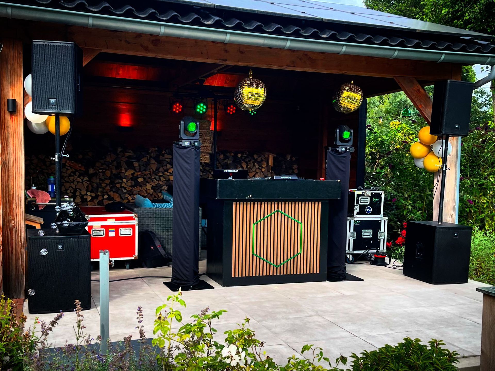 DJ-setup in een prieel met speakers, verlichting en een houten gevel. Gele ballonnen en brandhout op de achtergrond.