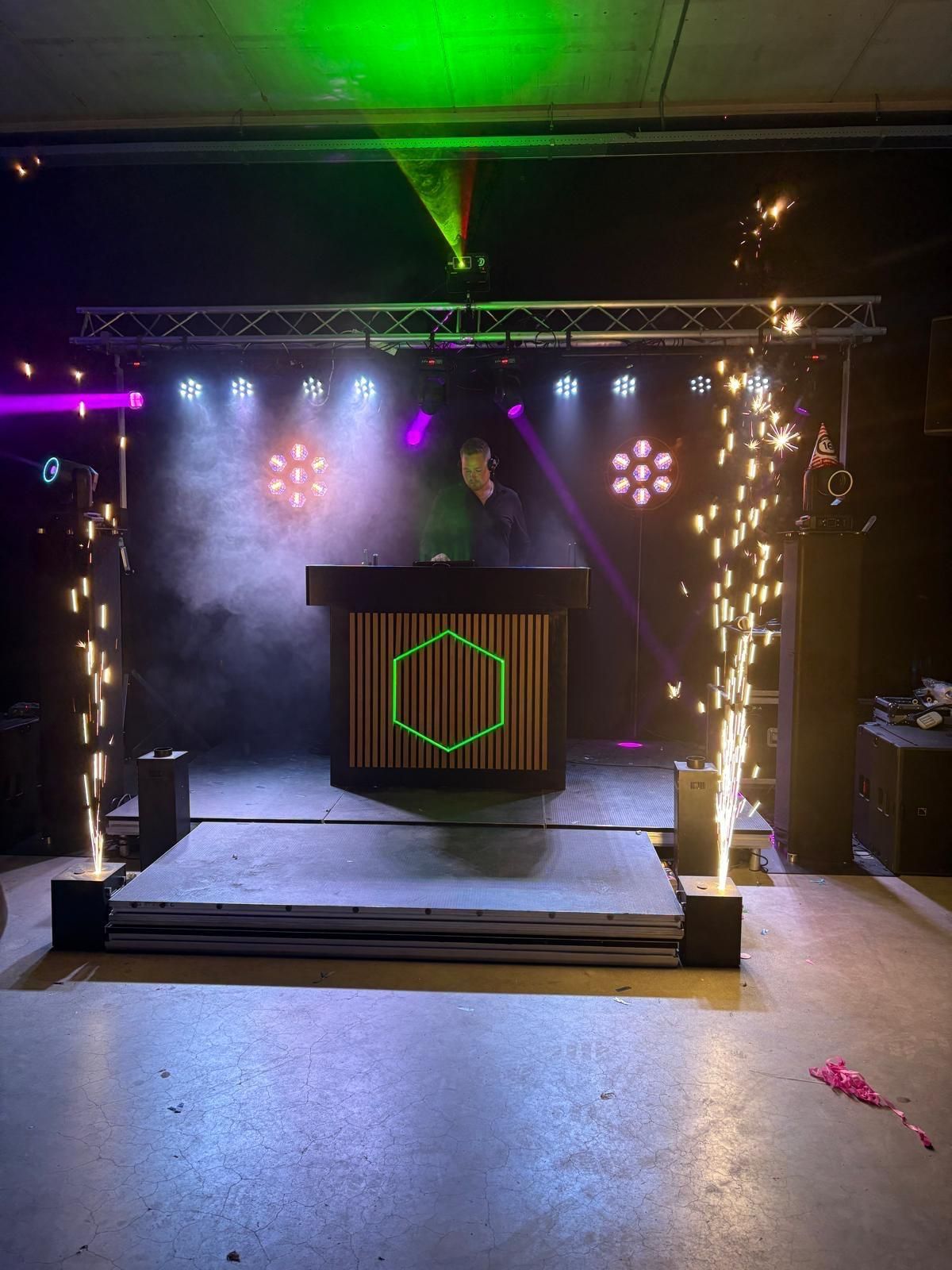 DJ in een club met podiumverlichting, rook en vonken. Zwarte muren.