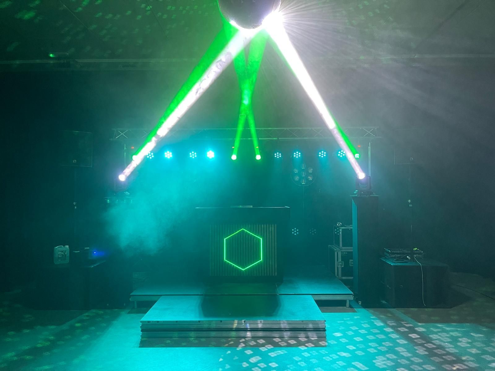 Podium met groene en witte verlichting, een DJ-booth met een oplichtende zeshoek en een spiegelbol.