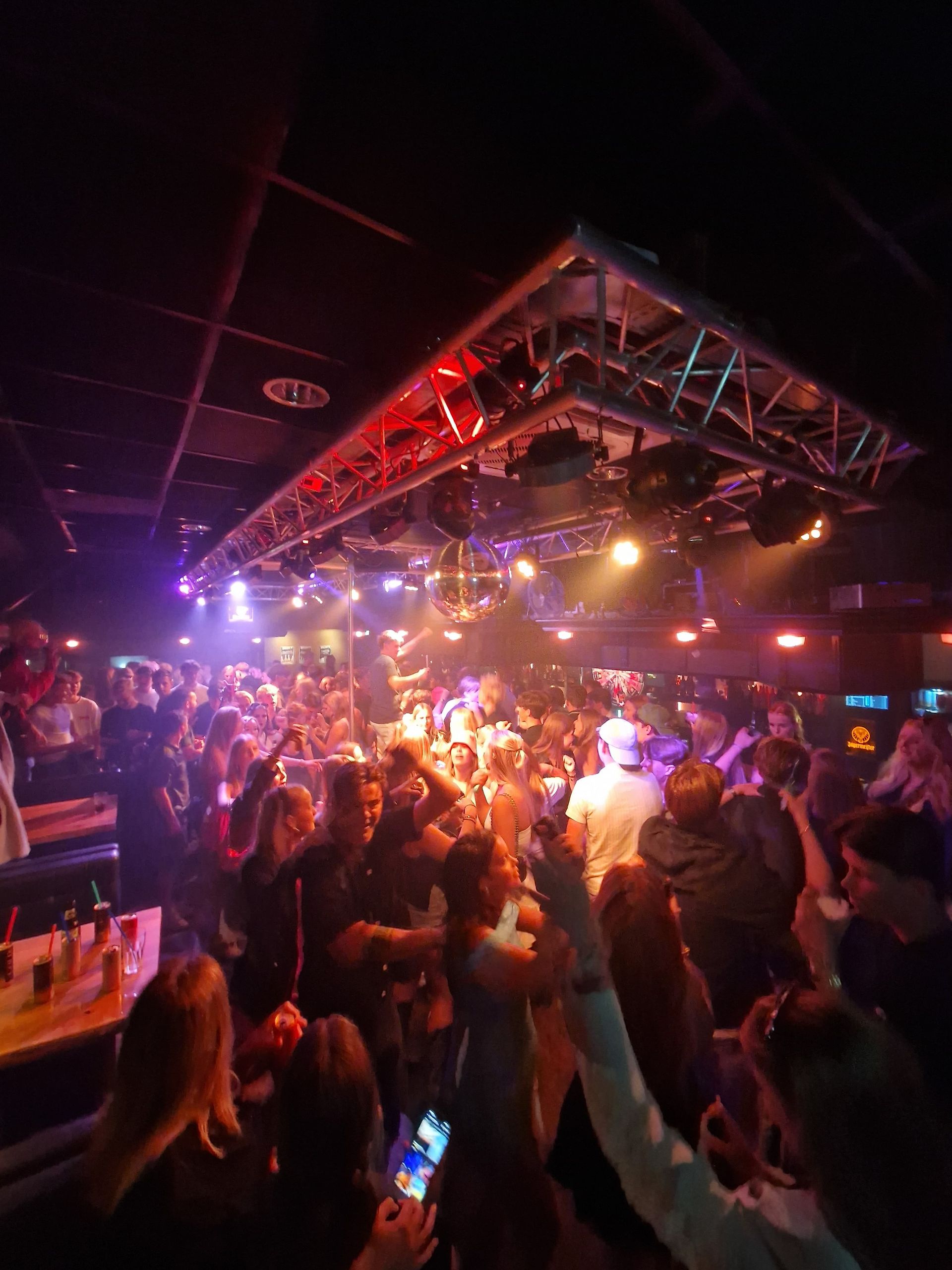 Mensen dansen in een drukke, schemerige nachtclub met kleurrijke lampjes en een podium.