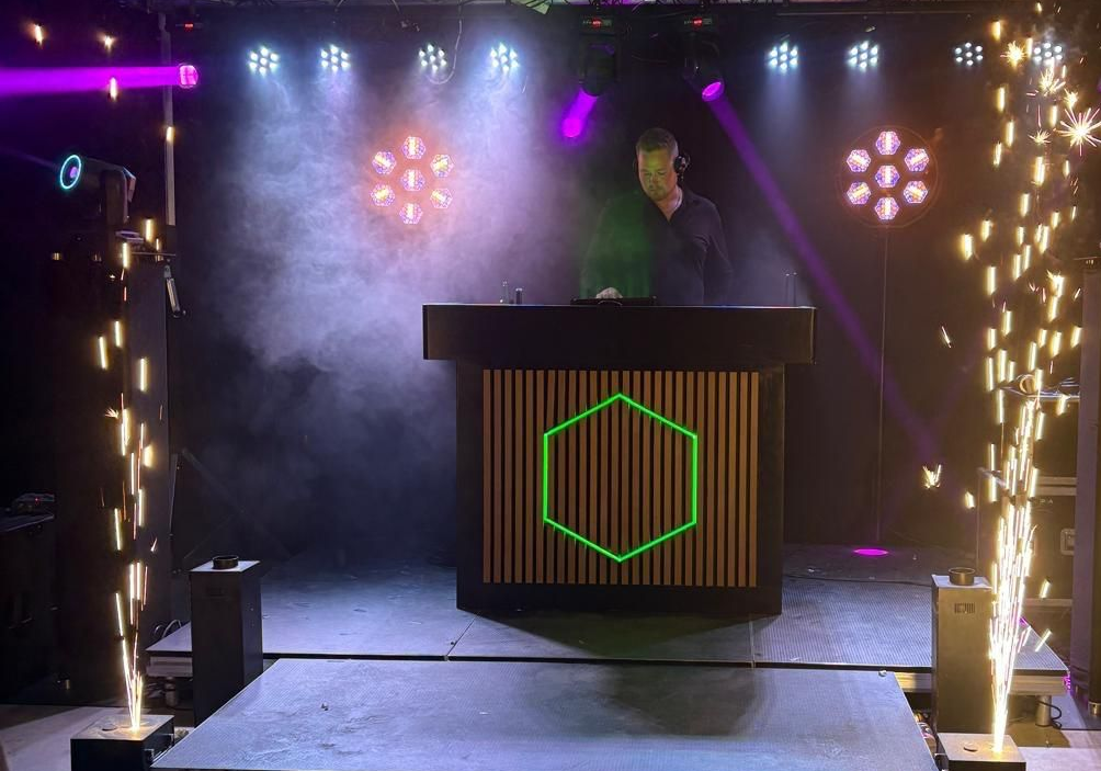 DJ die optreedt in een club met podiumverlichting, rook en sterretjes. De dj staat achter een zeshoekige dj-booth.