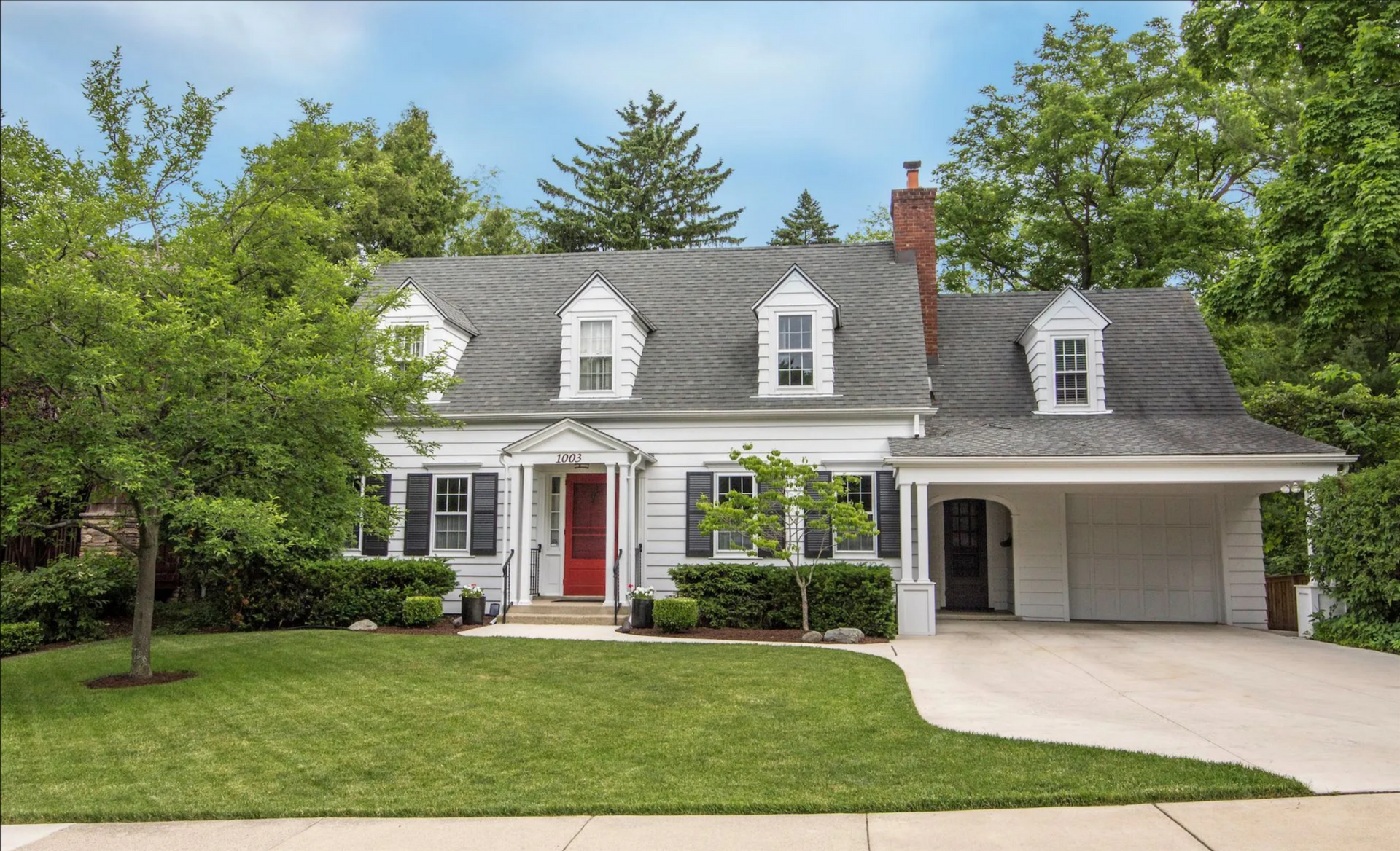 Suburban white house with gray roof, red front door, garage, and green lawn under trees