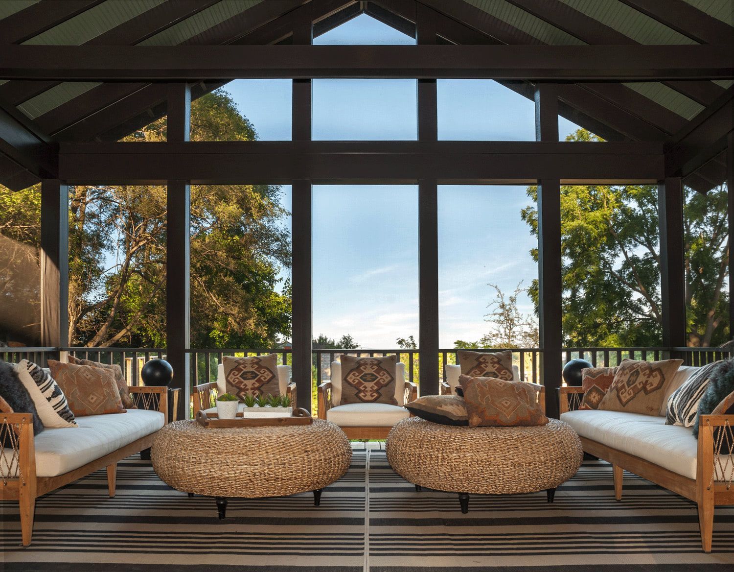 Cozy covered patio with striped sofas, woven ottomans, and large windows overlooking trees and blue sky
