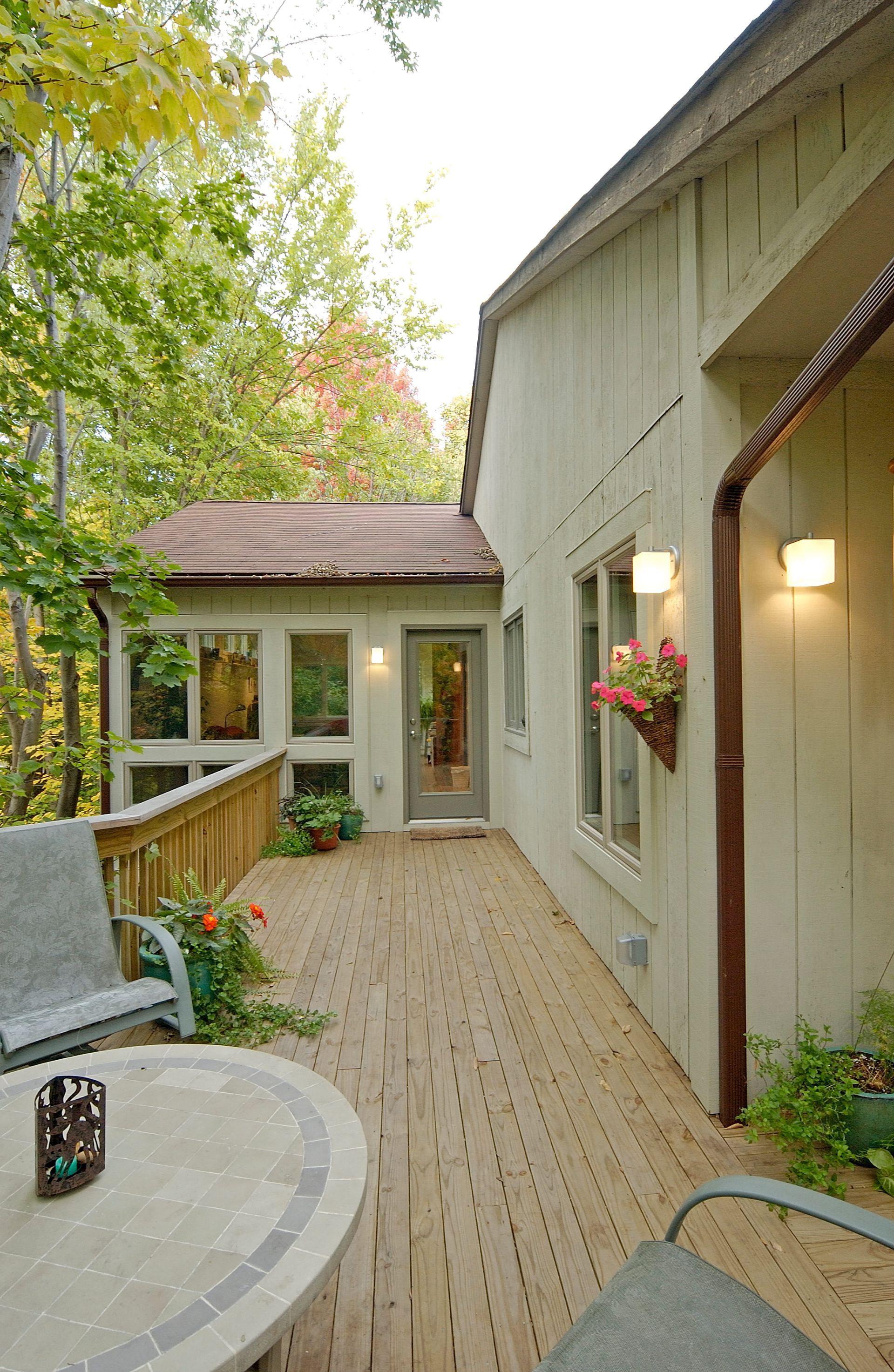 A wooden deck along the back of a house leading to a door and room with a wall of windows.