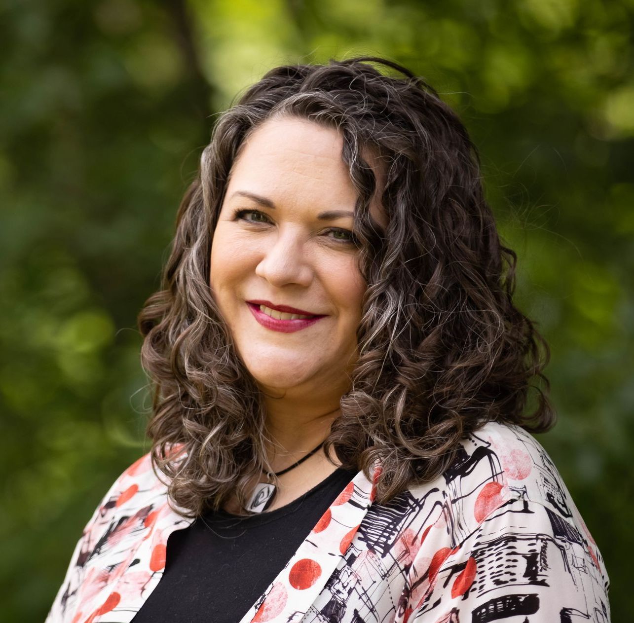 A woman with curly hair is smiling and wearing a floral jacket.