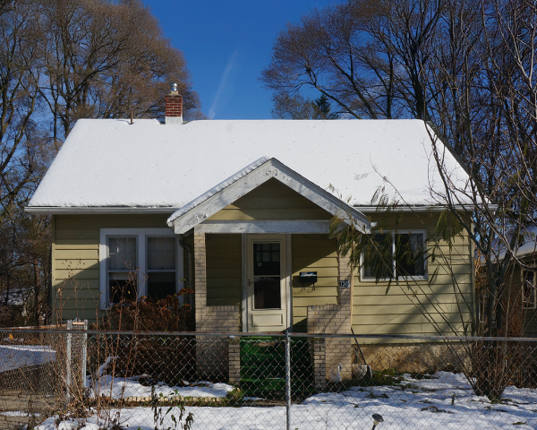 A small house with snow on the roof is behind a chain link fence