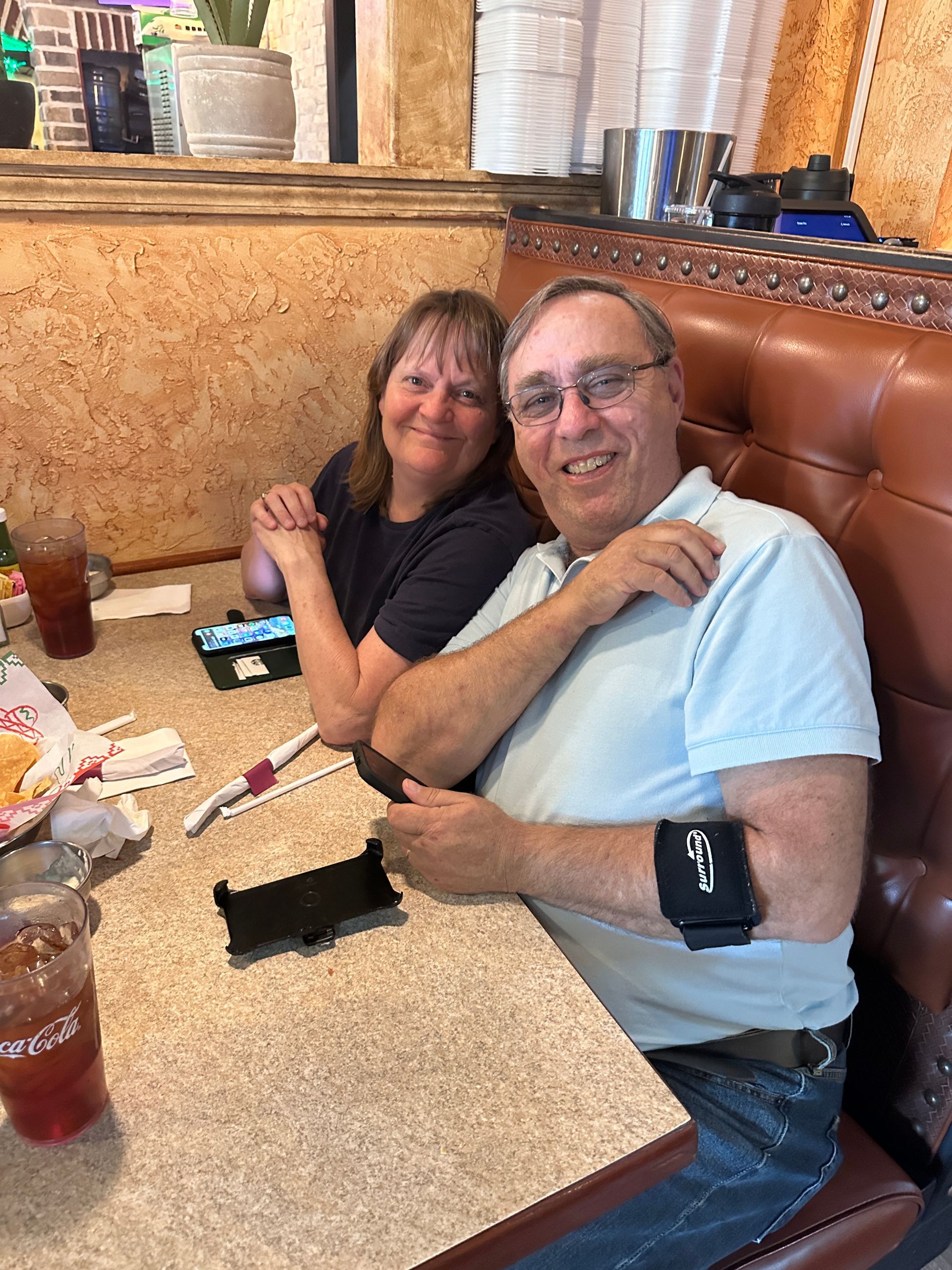 Couple at a restaurant table, smiling. Man has blood pressure monitor on his arm. Brown booth, tan wall.