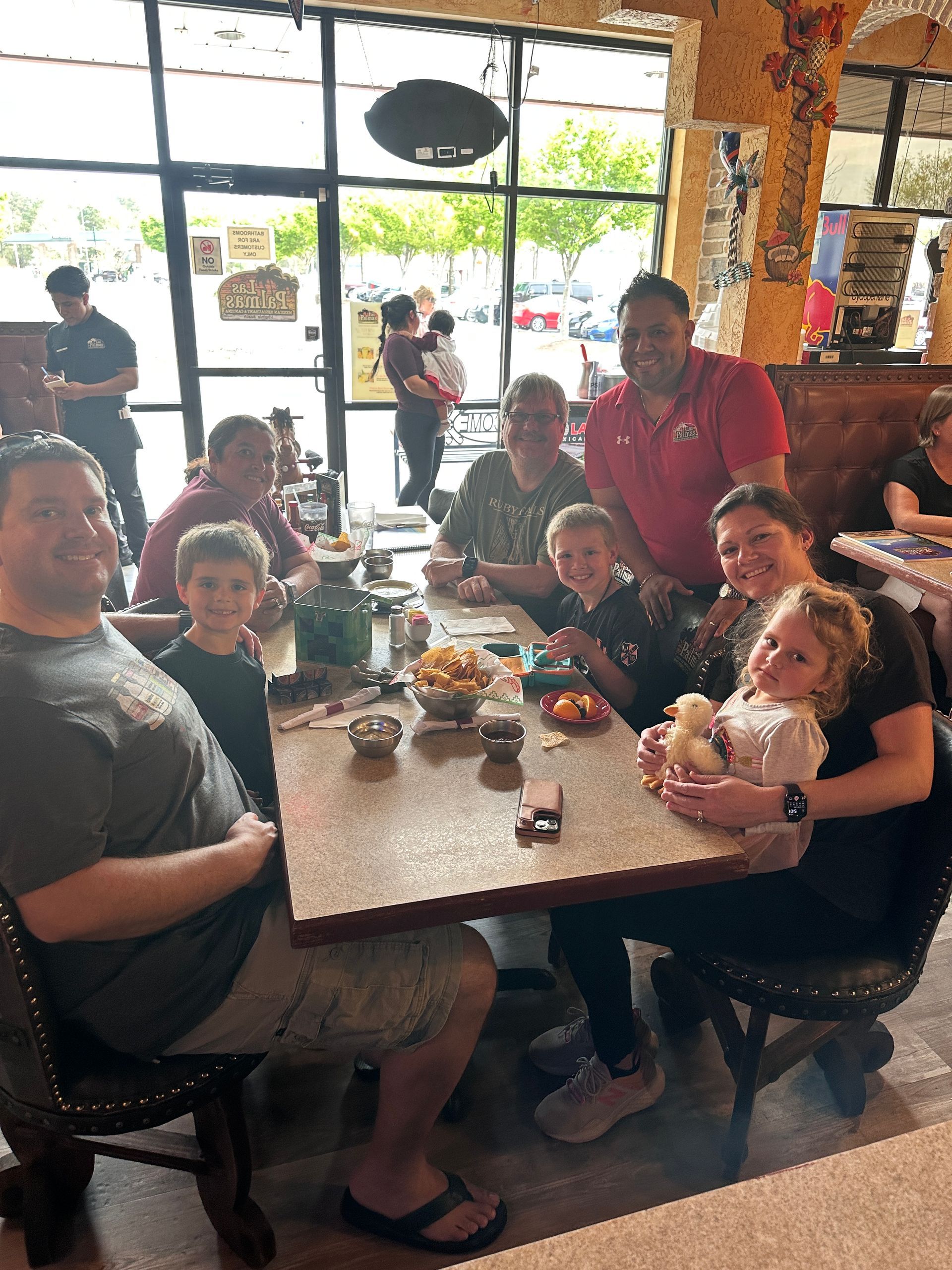 People sitting around a table in a restaurant; some are smiling at the camera. Sunlight through a large window.