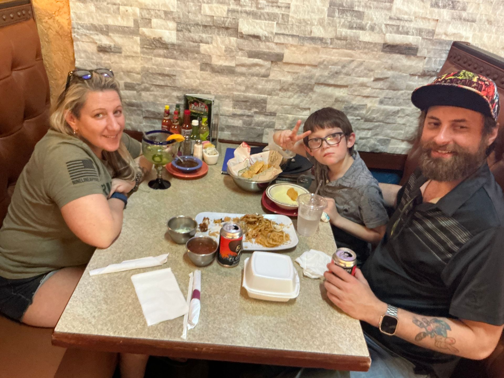 Family at a restaurant table with food. Woman smiling, boy making a peace sign, and man with a beard holding a can.