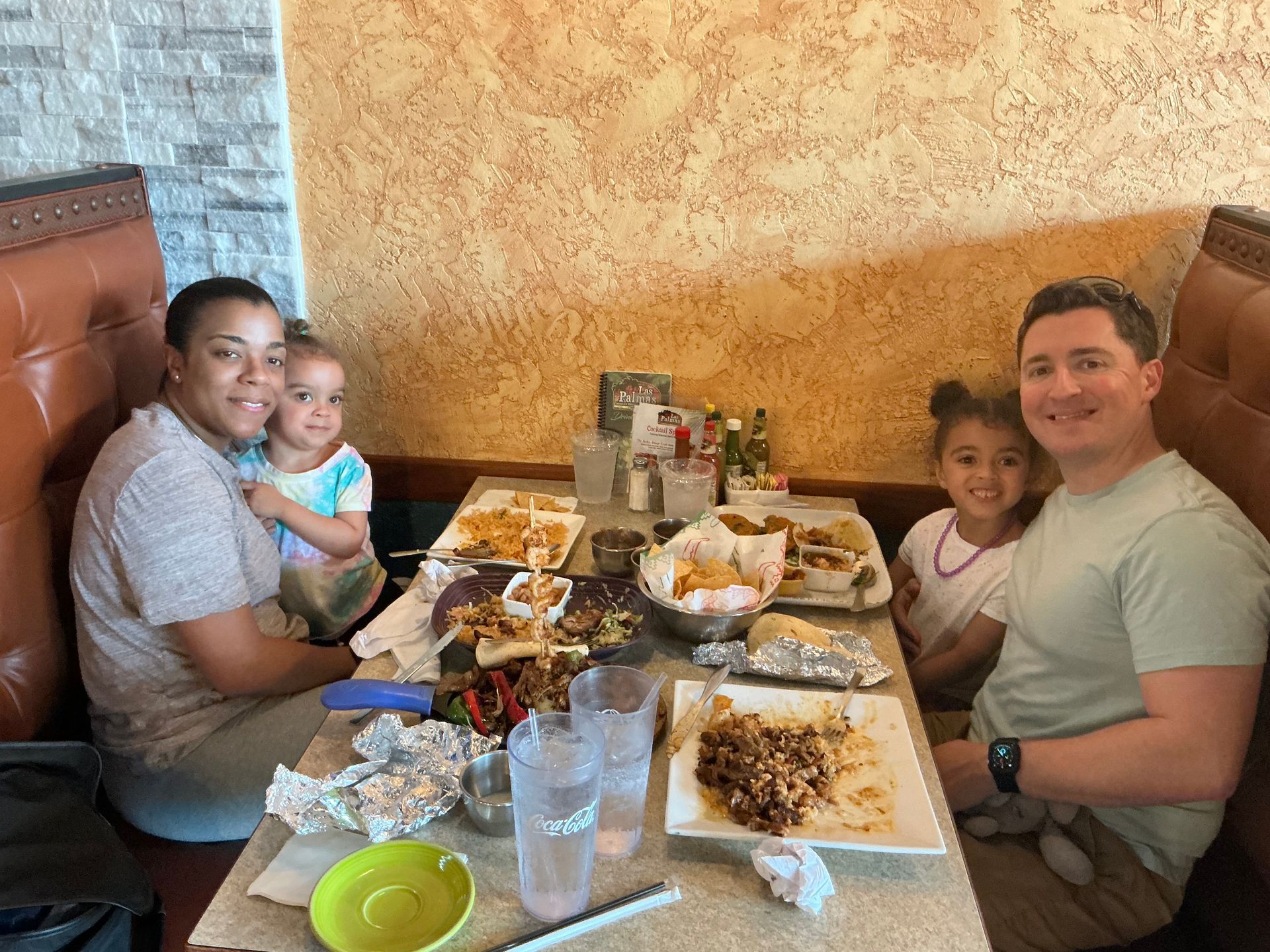 Family seated at a restaurant table with food; two adults and two children smile.