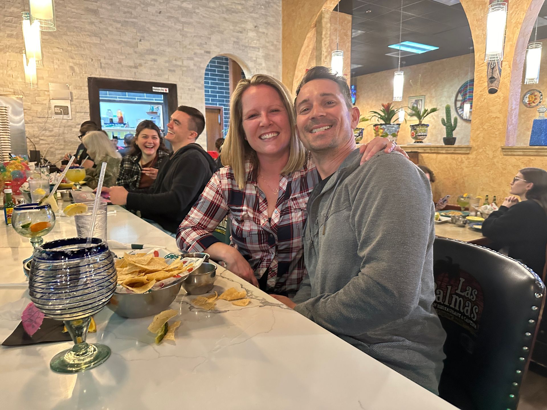 Couple smiling, arms around each other at a bar in a Mexican restaurant. Food and drinks visible.