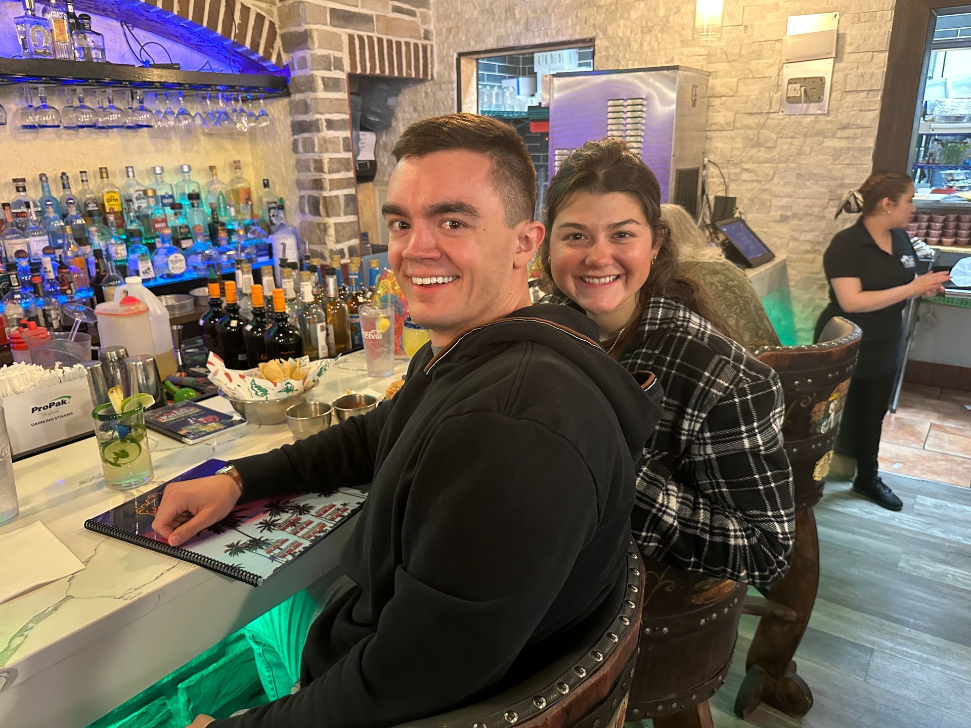 People smiling at a bar, man in a black hoodie, woman in plaid, bartender in the background.