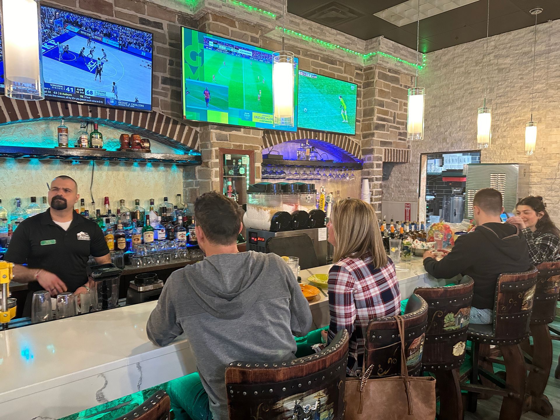 Bar with patrons and bartender; televisions display sports; interior setting.
