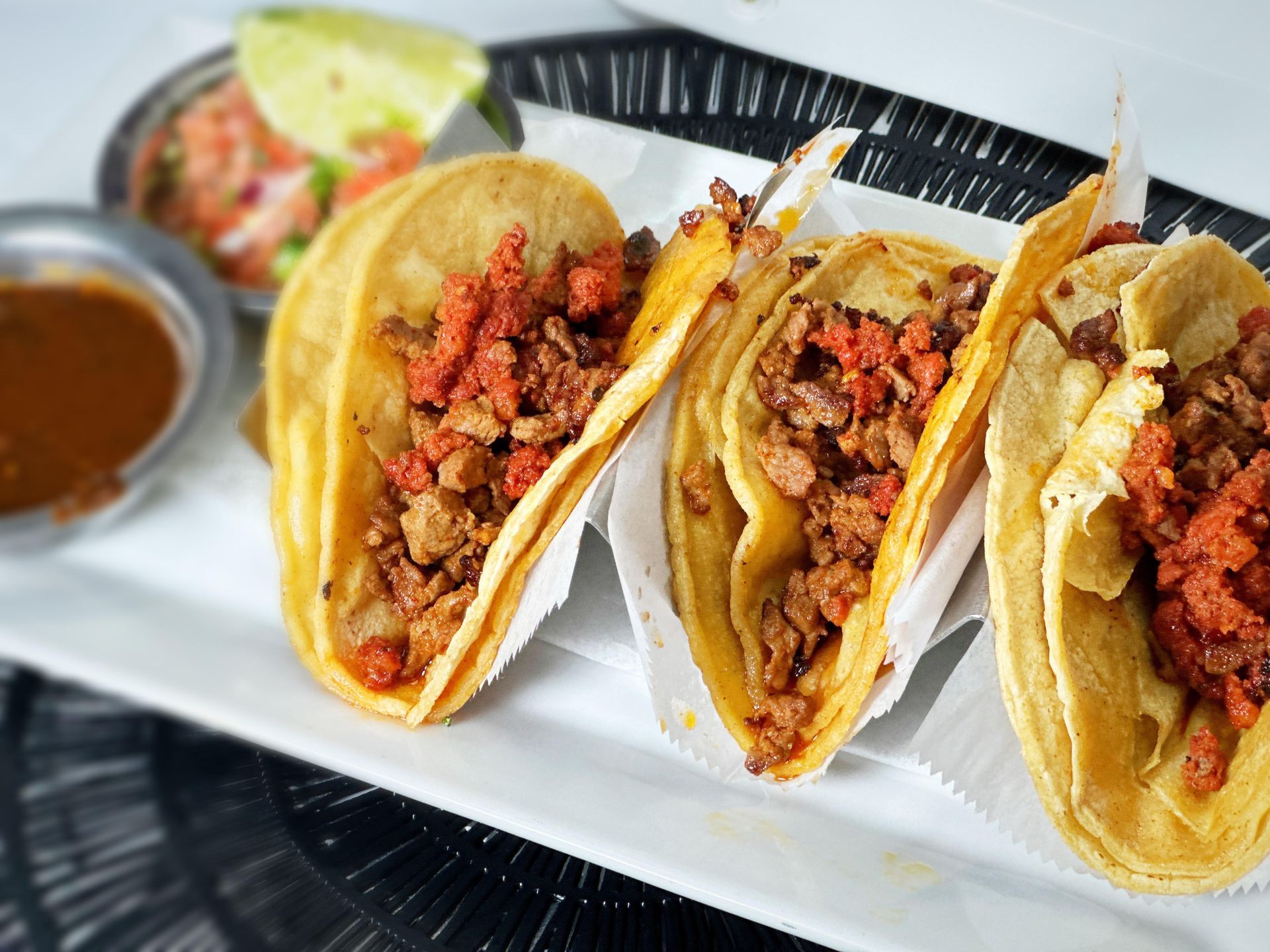 Three tacos on a white plate with salsa, lime, and sauce.