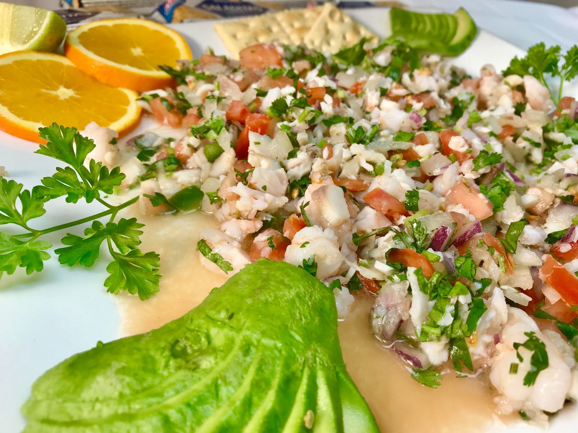 Ceviche with avocado, orange slices, lime, and crackers on a white plate.