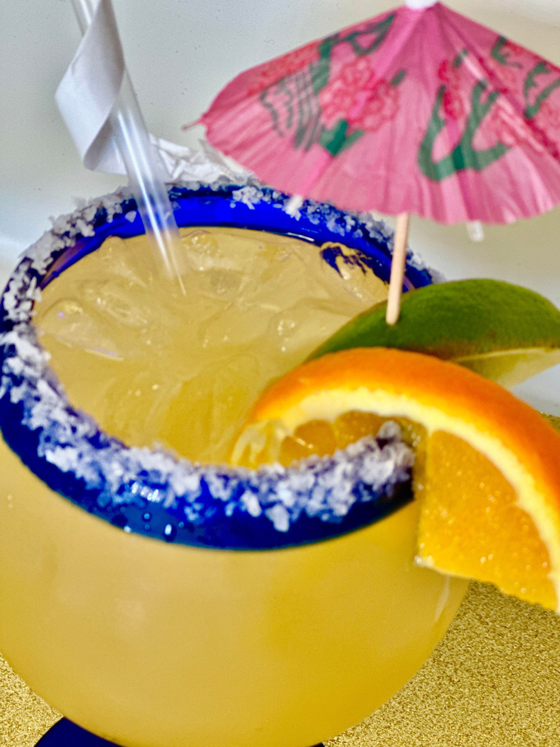 Margarita in blue-rimmed glass with salt, ice, straw, orange slice, lime, and pink umbrella.