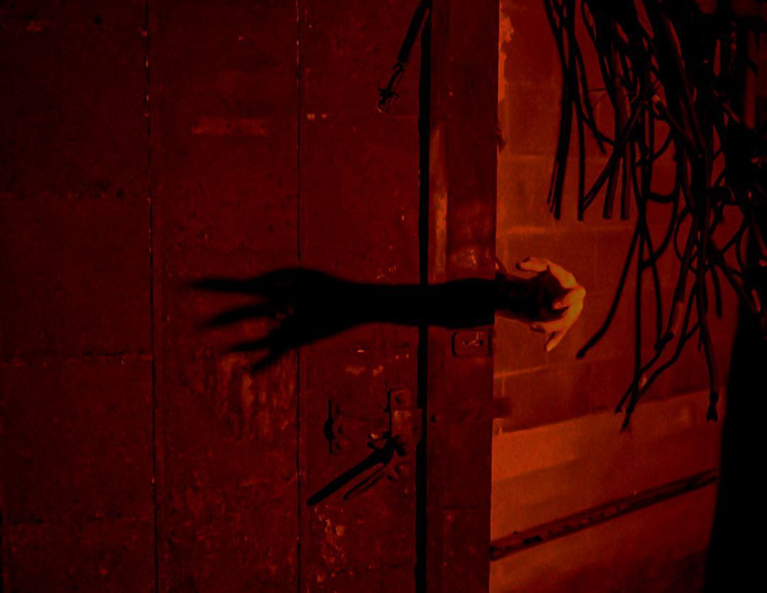 Shadowy hand reaching for a door handle in a dark red-lit room.