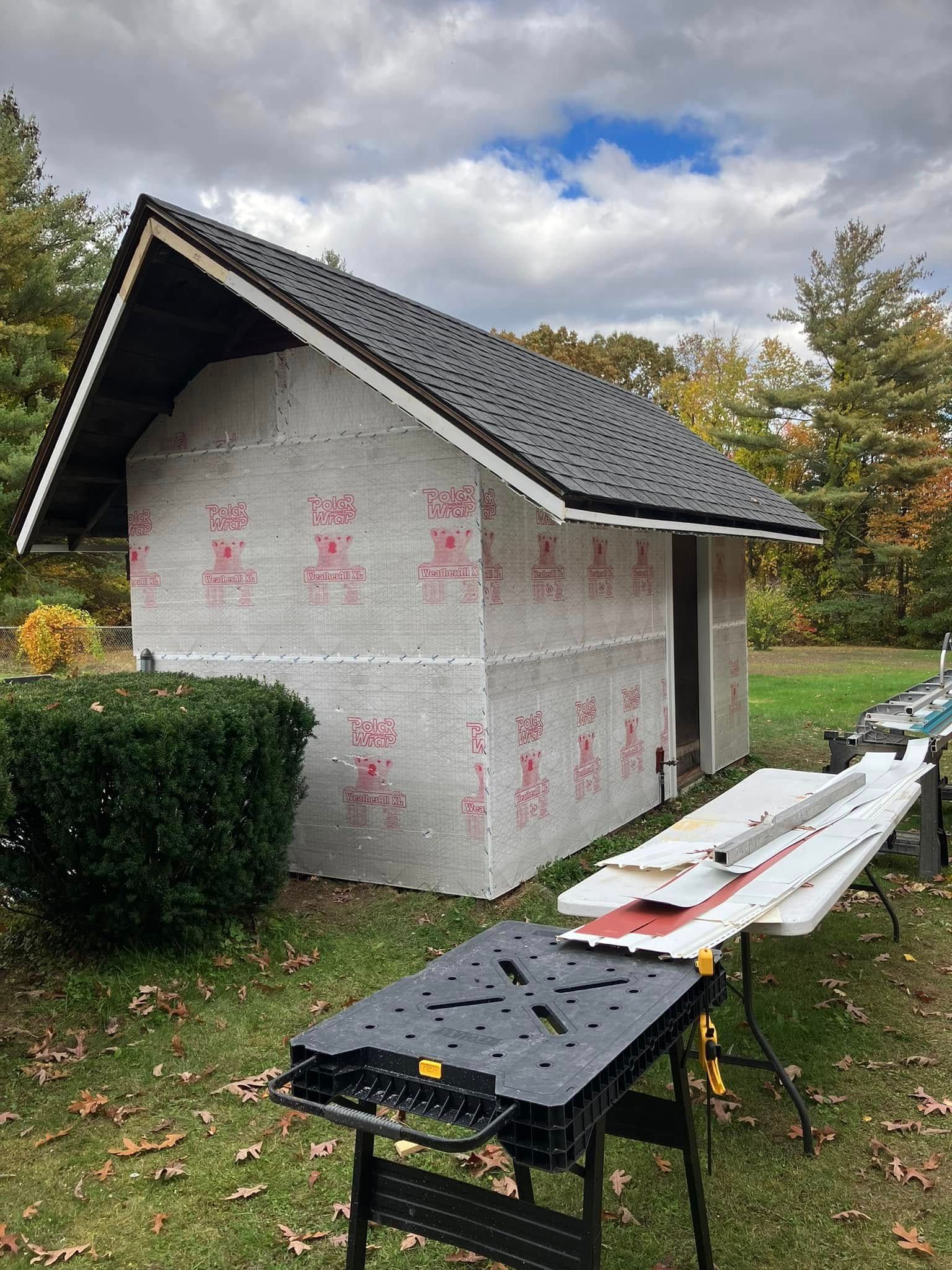 Small house under construction with white wrap, black roof, and workbenches outdoors on a grassy lot. Small house under construction with white wrap, black roof, and workbenches outdoors on a grassy lot.