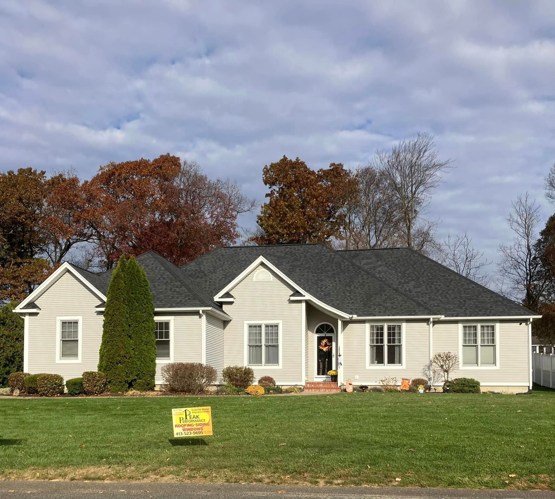 Single-story beige house with dark roof, front lawn, autumn trees, and cloudy sky Single-story beige house with dark roof, front lawn, autumn trees, and cloudy sky