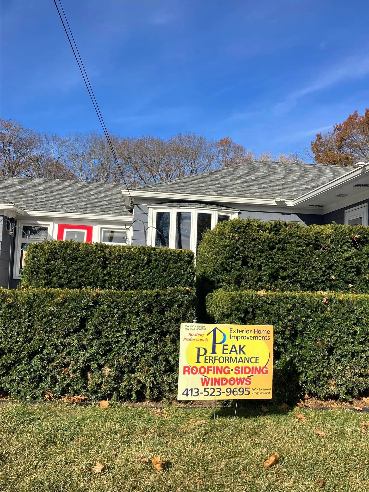 Yellow lawn sign in front of a house, reading Peak Roofing with phone numbers.