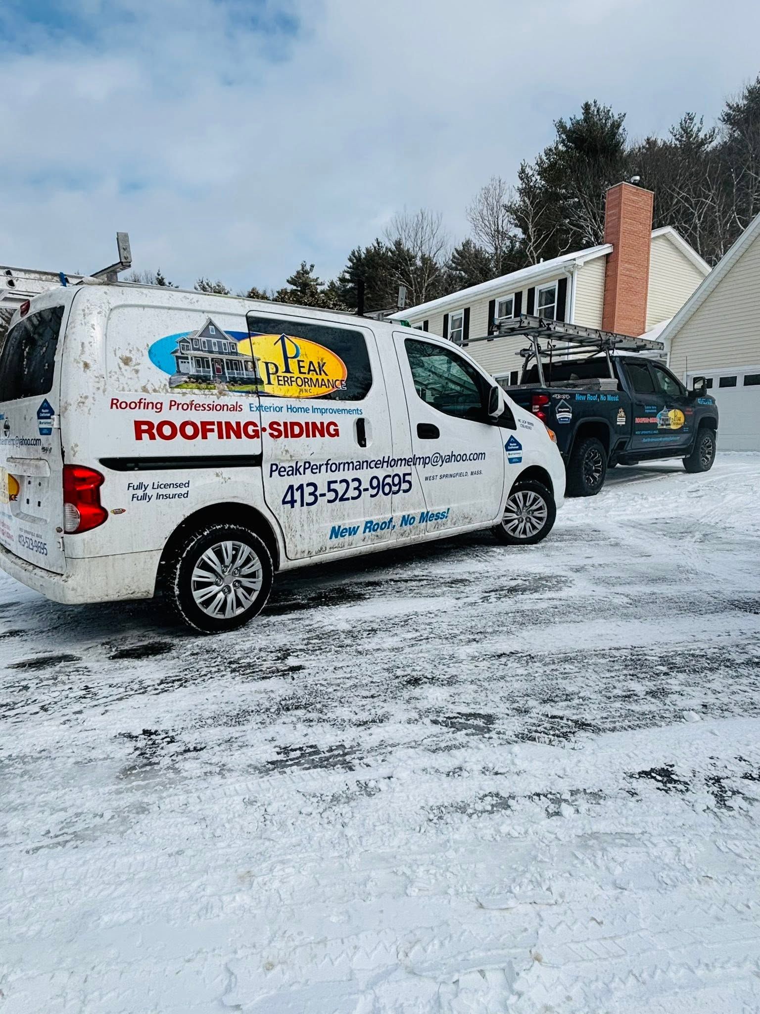 White roofing company van and black car parked in a snowy driveway by a house roof