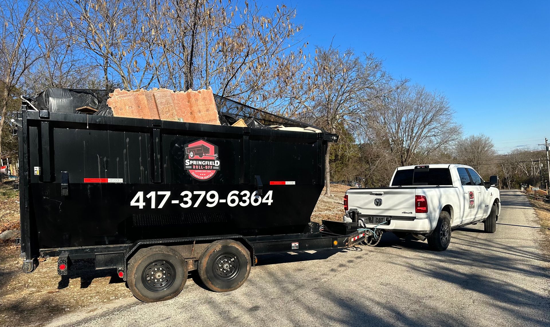 A dumpster is being towed by a truck on a dirt road.
