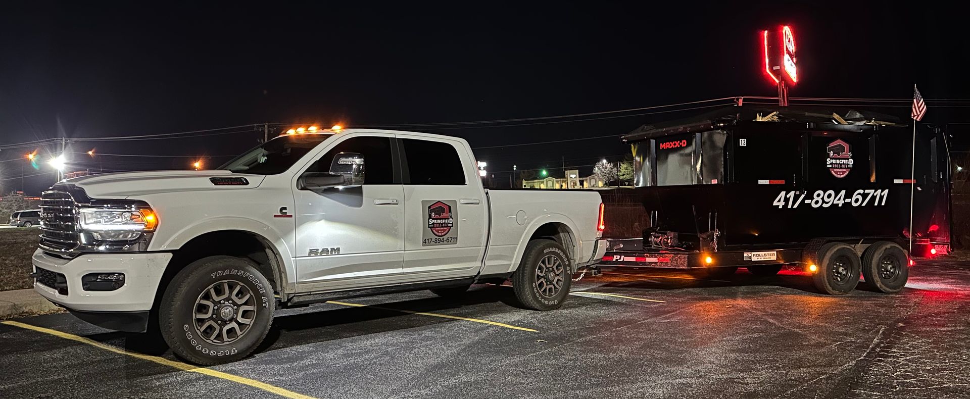 A white truck is towing a black trailer in a parking lot at night.