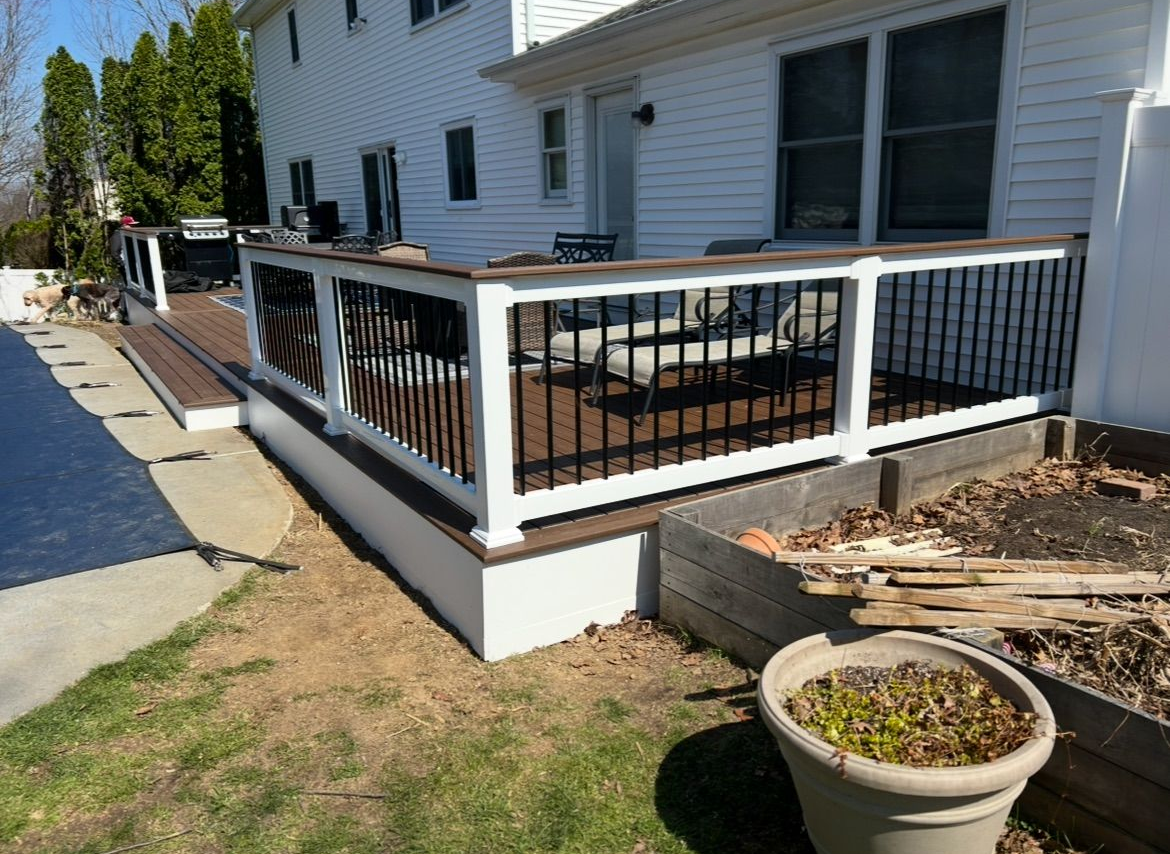 White house deck with black railing, patio table, and potted plants beside a sidewalk