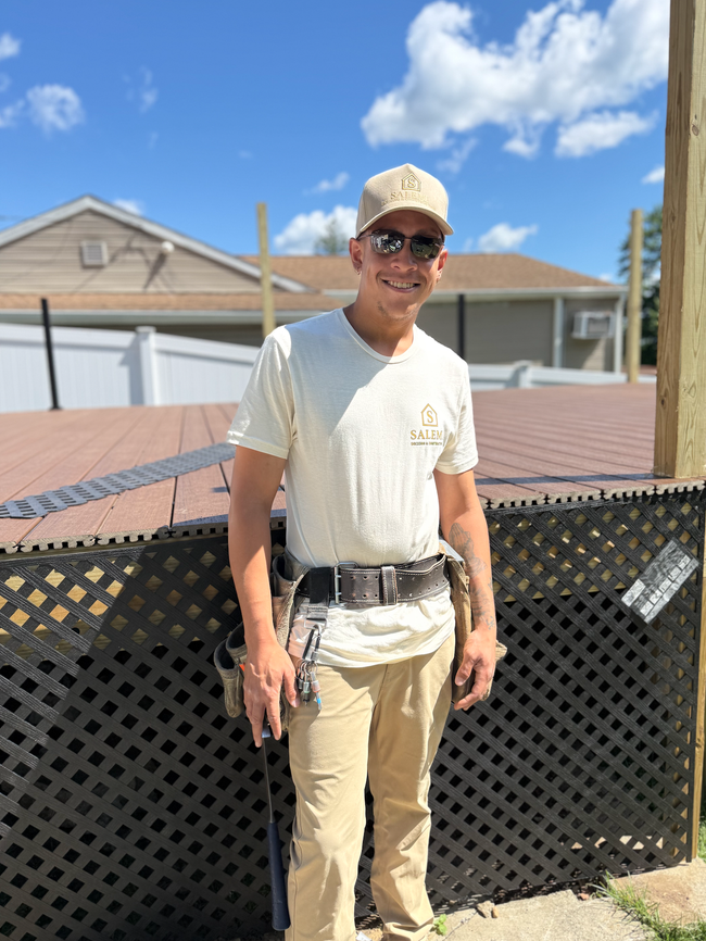 Person standing outdoors by a fence in a tan shirt and cap, smiling under a blue sky.