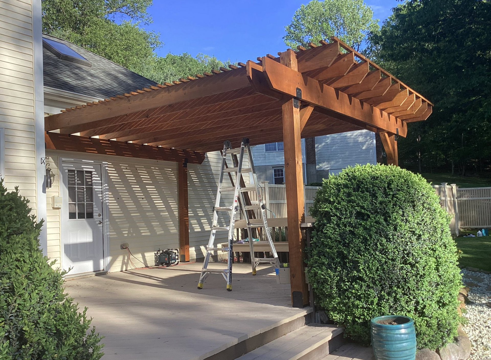 Wooden pergola under construction in a backyard, with ladders and trimmed shrubs nearby.