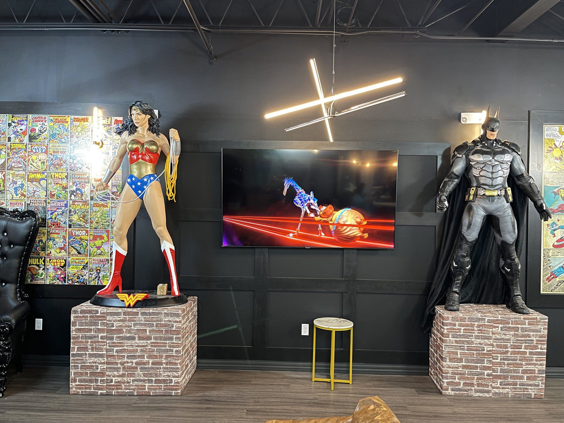 Batman and Wonder Woman statues flanking a TV display in a dark gallery with neon cross lights