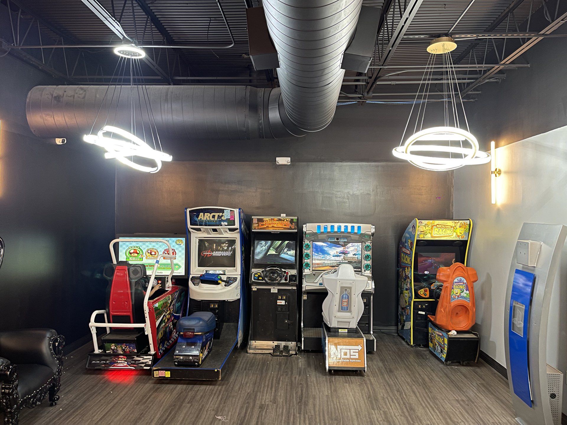 Arcade room with five arcade machines against a dark wall and overhead lights, including blue vending machine on right